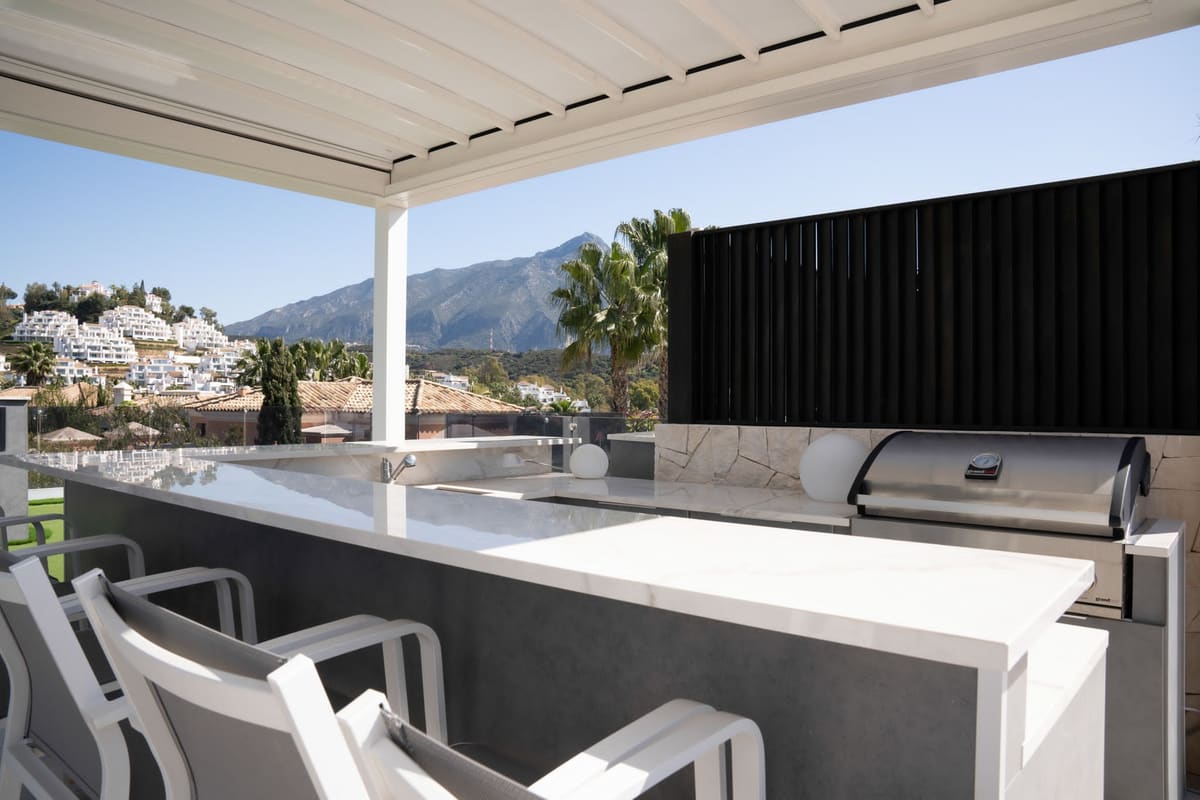 Outdoor kitchen bar with white worktop and built-in BBQ under a louvred pergola, with La Concha mountain behind