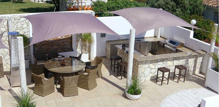 Stone-clad U-shaped outdoor kitchen with bar stools and a dining area under two sail shades