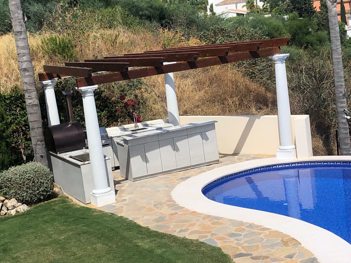 Wooden pergola on white columns over an outdoor kitchen with a smoker, beside a garden pool