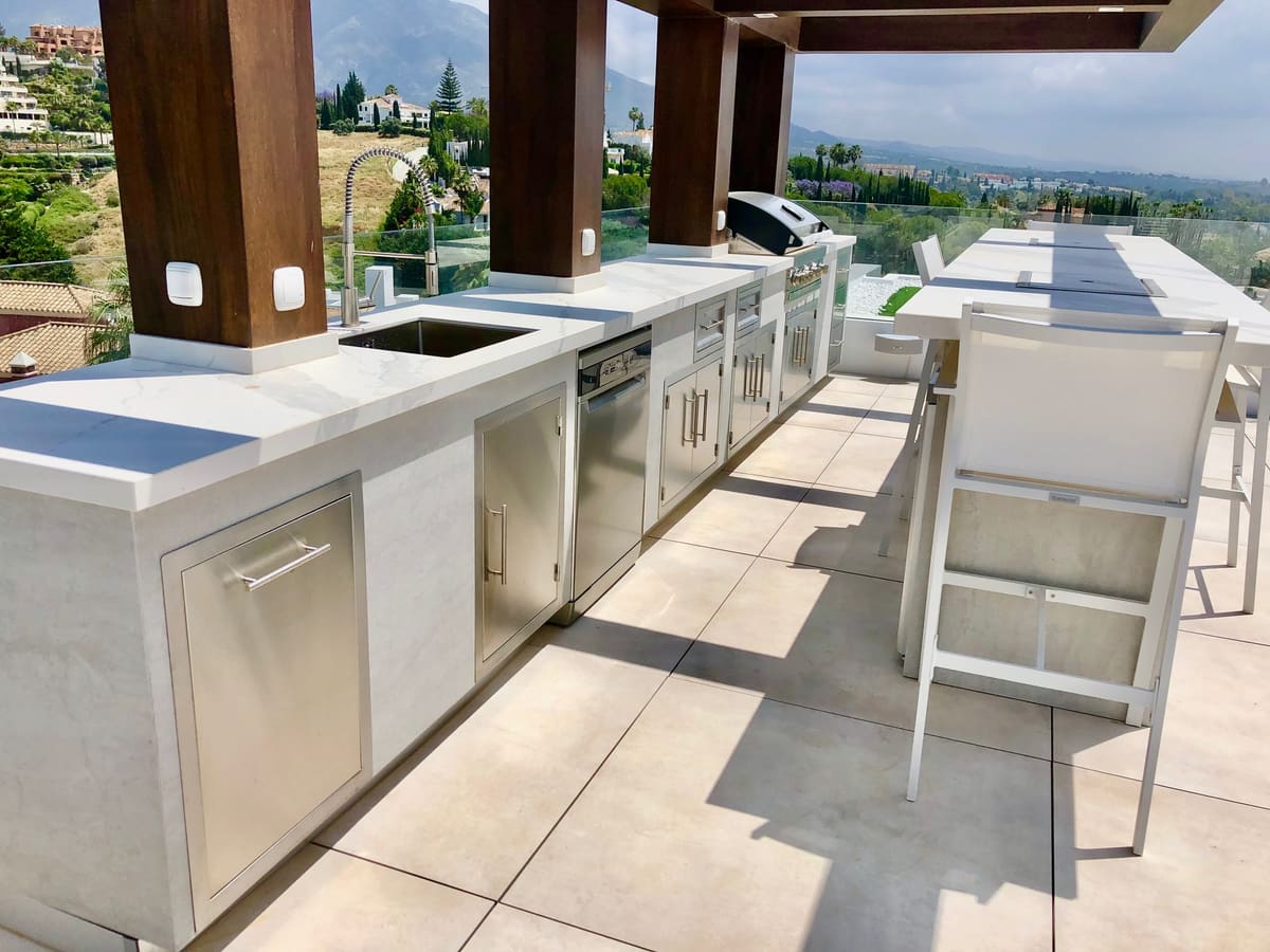Built-in outdoor kitchen with stainless steel doors, sink and BBQ under timber beams on a terrace with mountain views