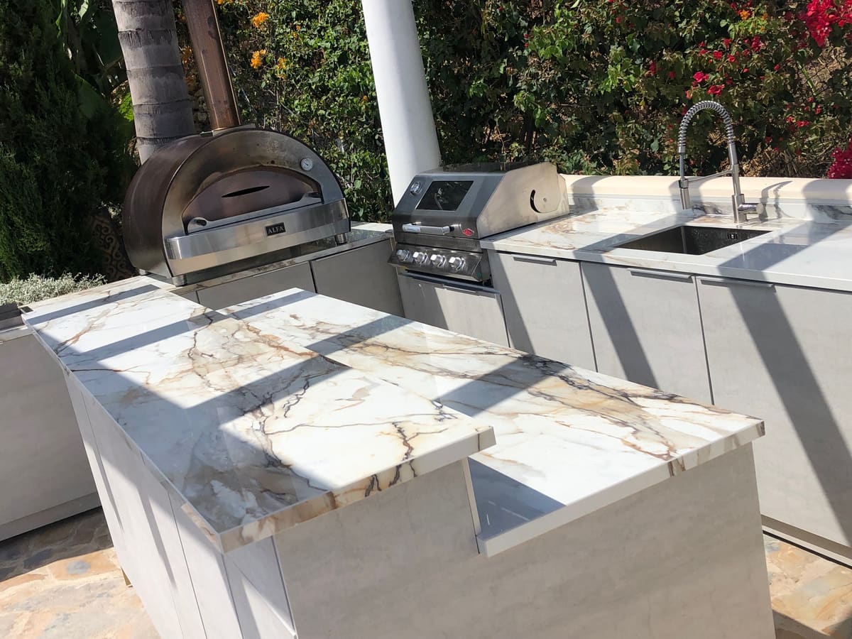 Stainless steel pizza oven beside a built-in gas BBQ and a marble-effect worktop in a garden kitchen with bougainvillea