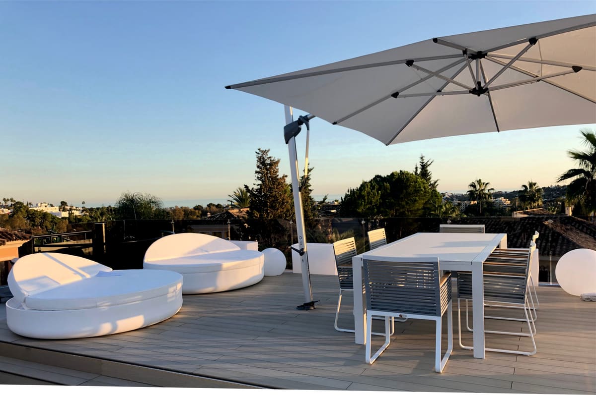 Large cantilever umbrella over a dining table and daybeds on a decked terrace at dusk, with the sea on the horizon